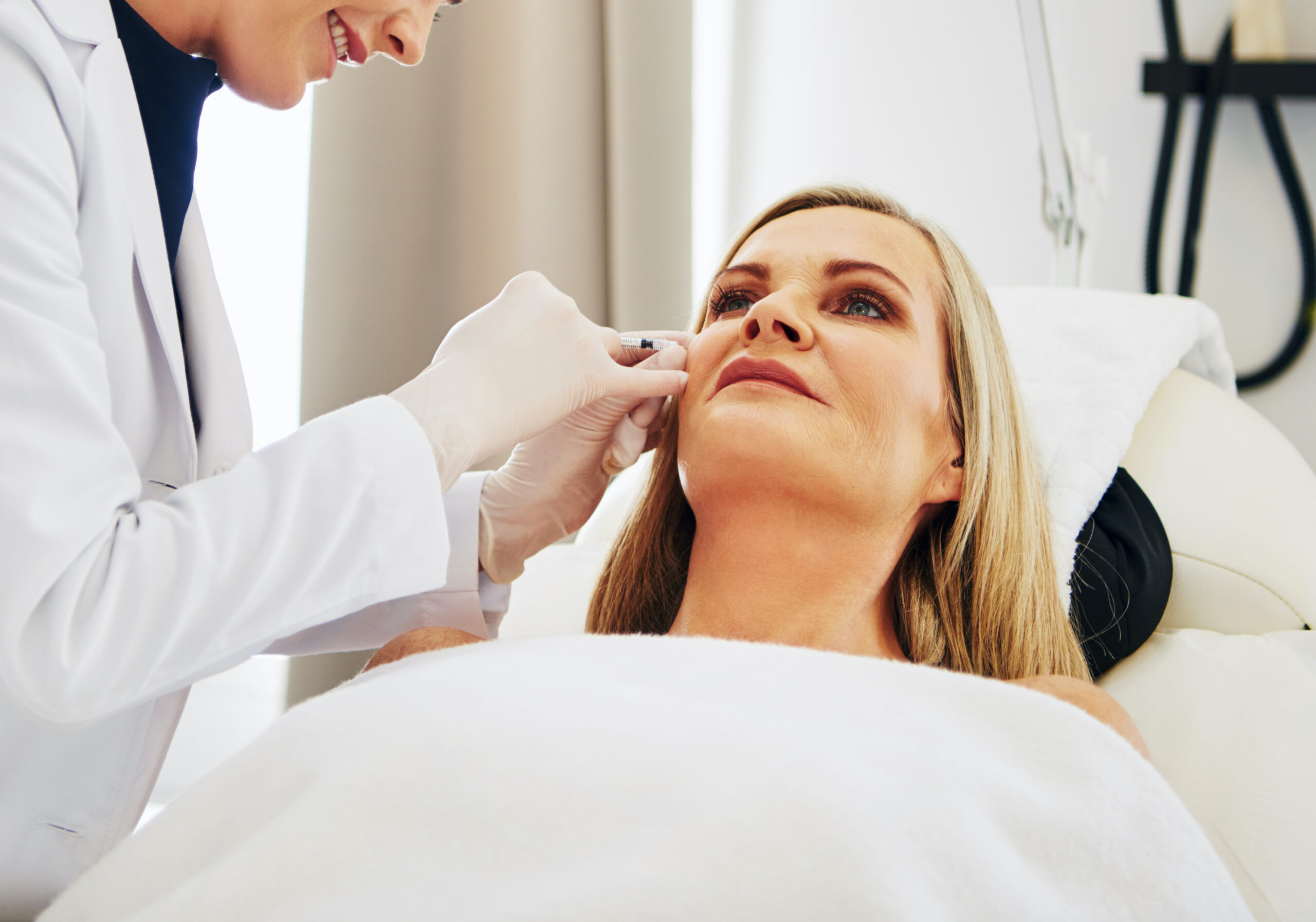 Smiling young female doctor doing botox injections on the face of a mature woman lying on a table in a beauty clinic